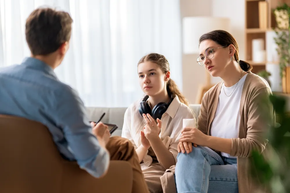A counselor taking notes on a clipboard during a session with a teenager and her mother