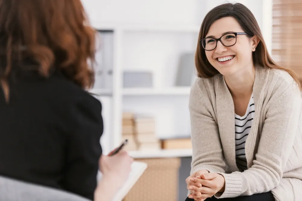 A woman in glasses and a cream cardigan smiles during a session while a therapist takes notes on a pad in the foreground