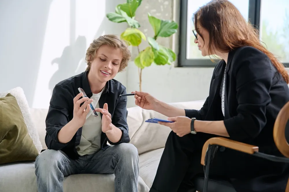 A therapist holding a clipboard and pen listening to a teenage client on a sofa