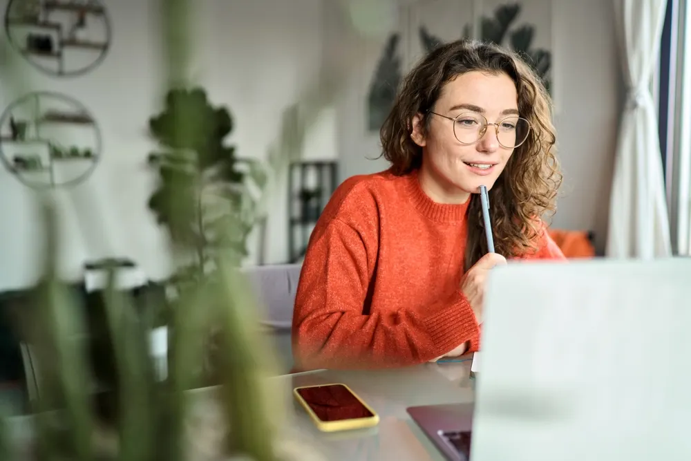 A student in an orange sweater and round glasses holds a pen to her chin while reading her laptop screen at a table