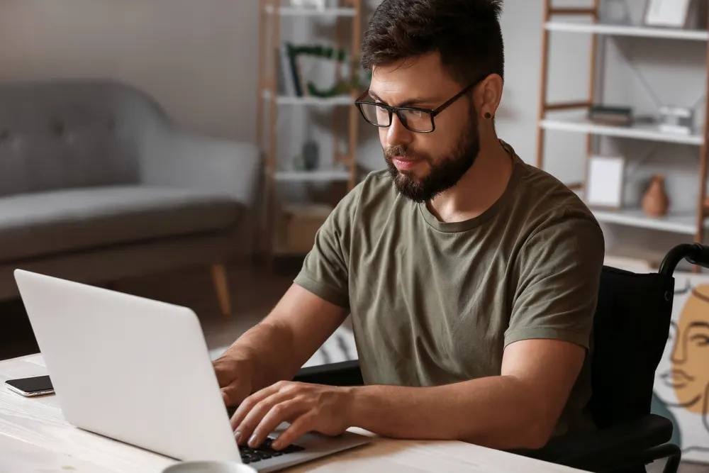 A man in a wheelchair working at a laptop at a desk in a home study
