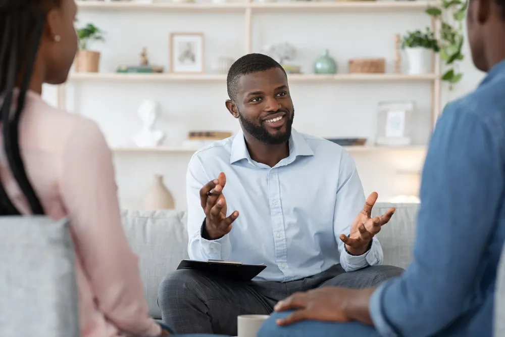 A counselor holding a clipboard gesturing while speaking with a couple during a session