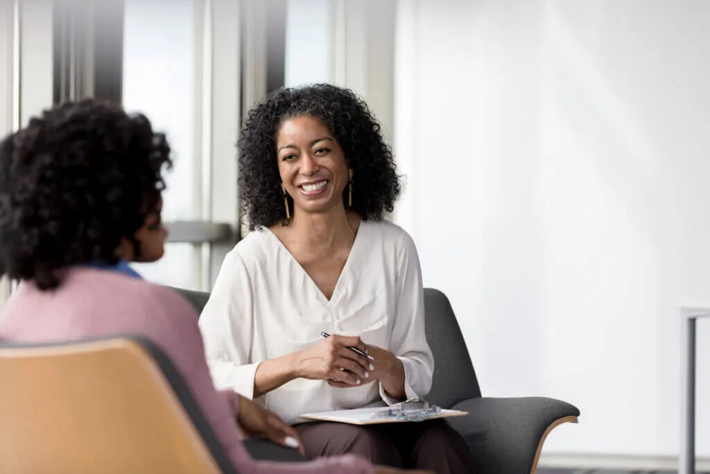 A smiling counselor in a white blouse holds a pen and clipboard while seated across from a client in a bright office