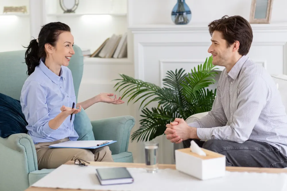 A therapist gesturing with a pen and clipboard while talking with a smiling client in a bright living room