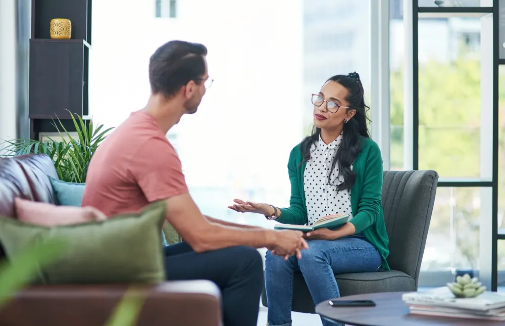 A counselor with an open notebook speaking with a client during a session in a sunlit office