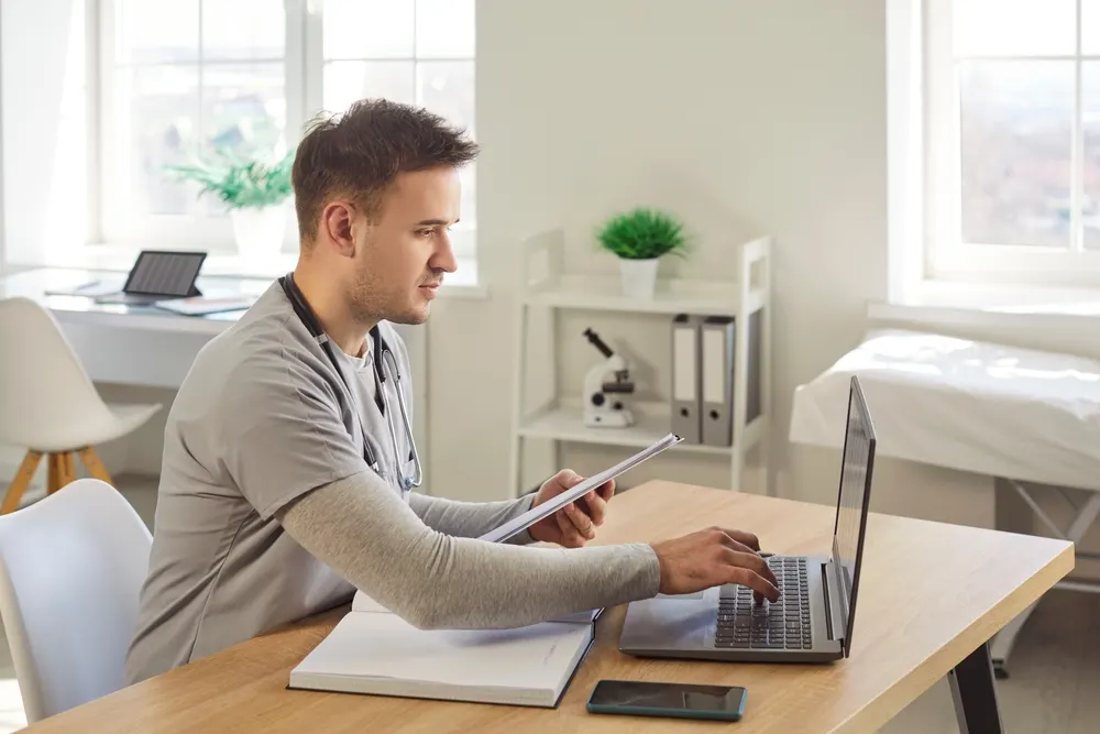 A healthcare worker in scrubs holding notes while typing at a laptop in a bright clinic office