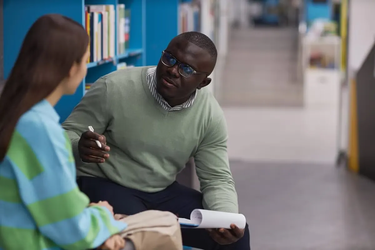 A school counselor holding papers and a pen talks with a student seated beside him in a school library