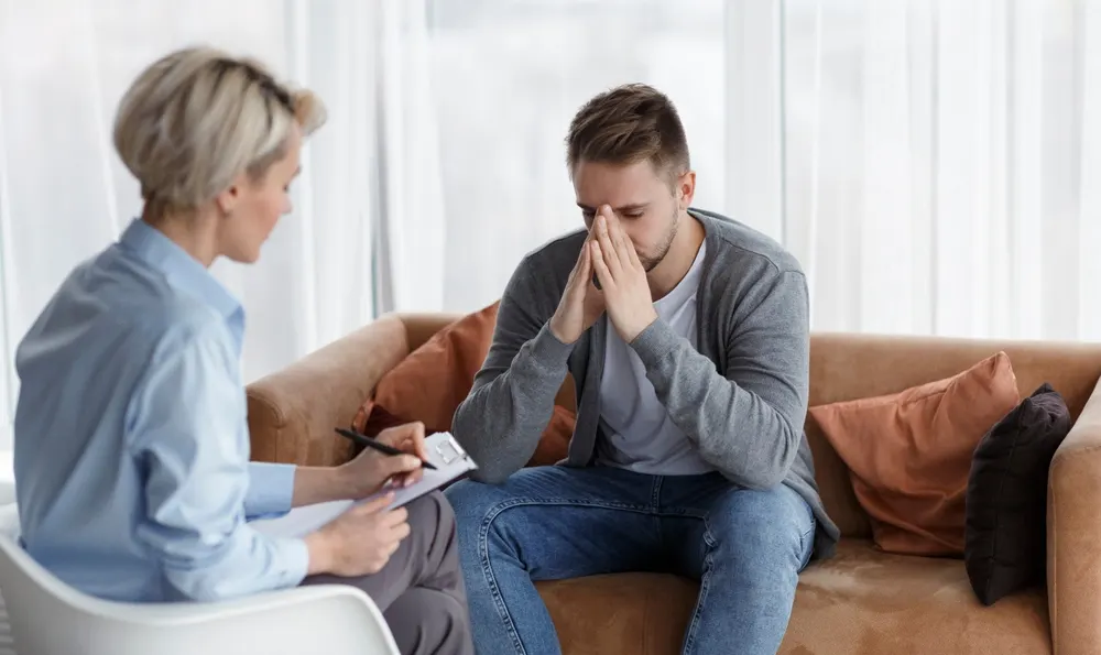A counselor taking notes while a distressed man sits with his hands to his face on a sofa